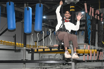 A young man on a zip line at an indoor adventure park, waving to the camera.