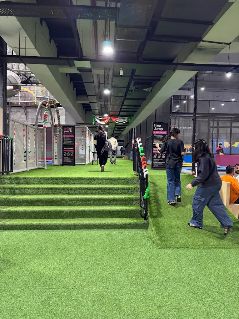 People walk through a brightly lit indoor trampoline park with green turf and colorful decorations.
