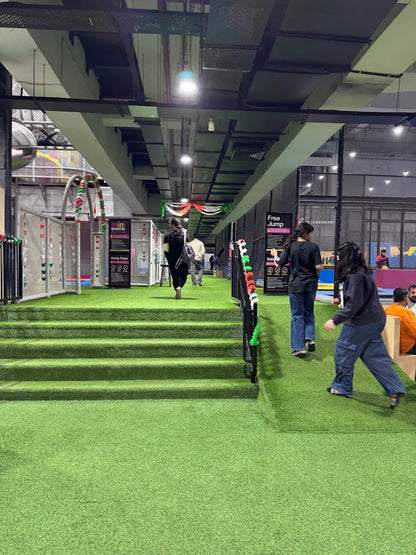 People walk through a brightly lit indoor trampoline park with green turf and colorful decorations.