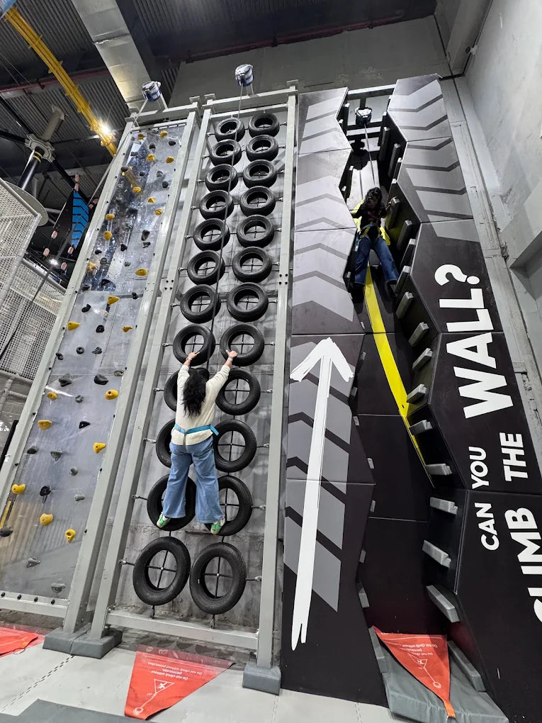 Two young people climb on tire and rock climbing walls in an indoor adventure park.