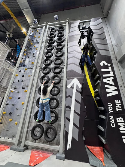 Two young people climb on tire and rock climbing walls in an indoor adventure park.