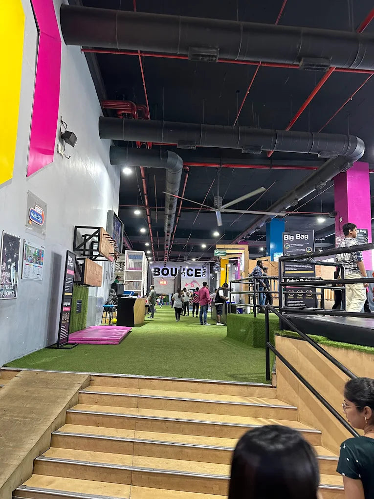 People walk through an indoor trampoline park with green turf and large pipes overhead.