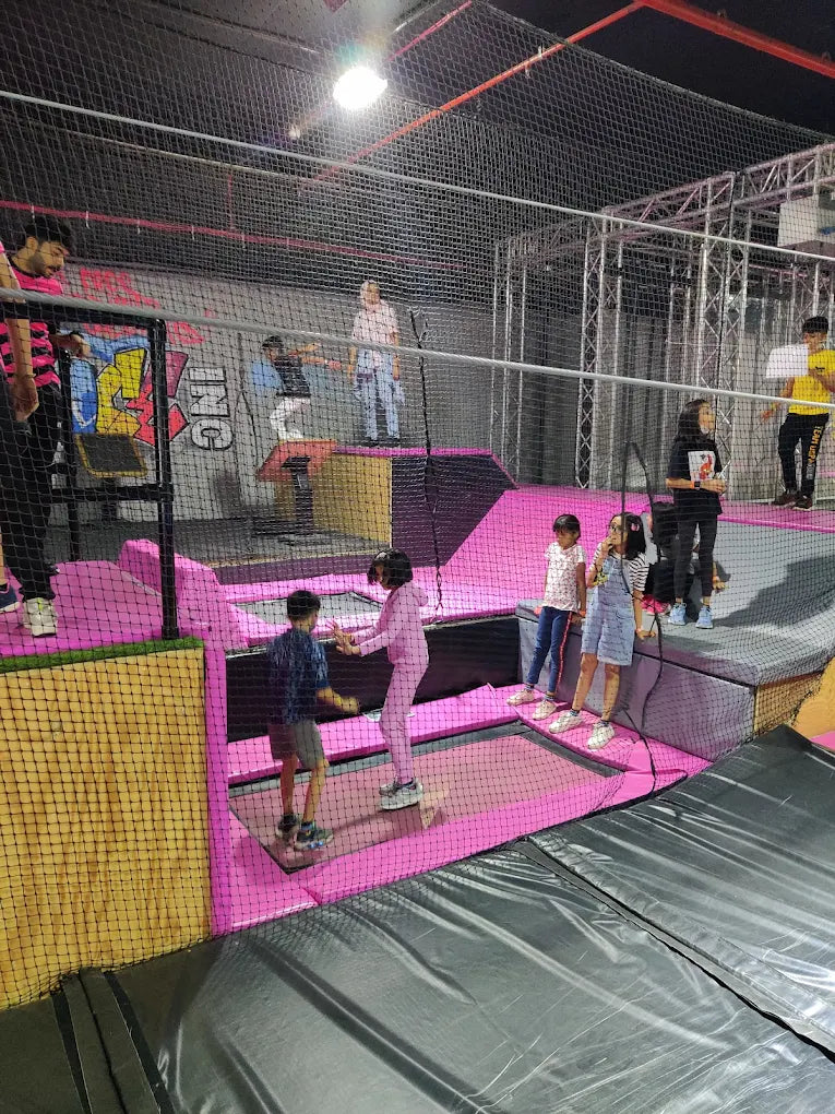 Children and adults enjoy a trampoline park with various interconnected trampolines and padded areas.