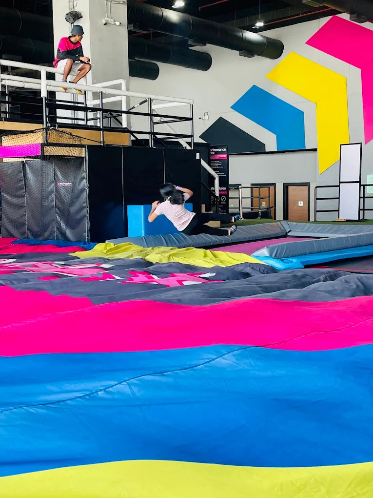 A young person jumps on a trampoline in an indoor park with colorful walls and mats.