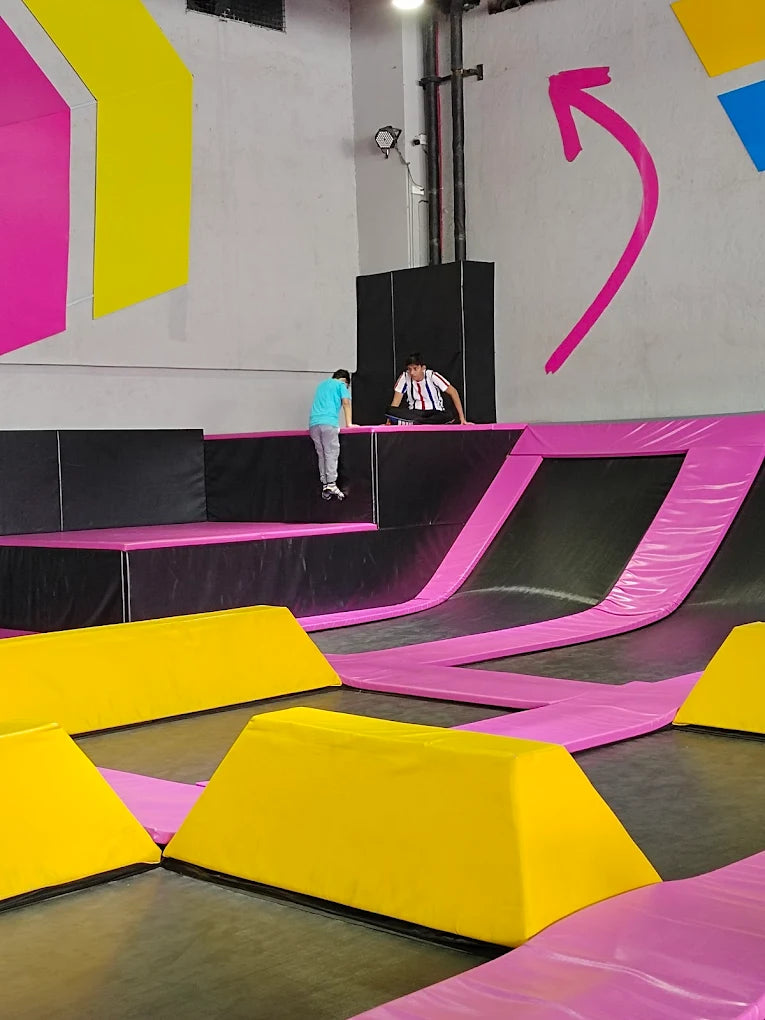 Two young people are playing on trampolines in an indoor trampoline park with colorful walls and padding.