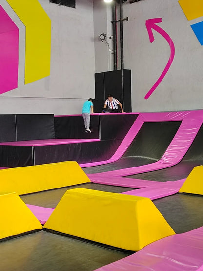 Two young people are playing on trampolines in an indoor trampoline park with colorful walls and padding.