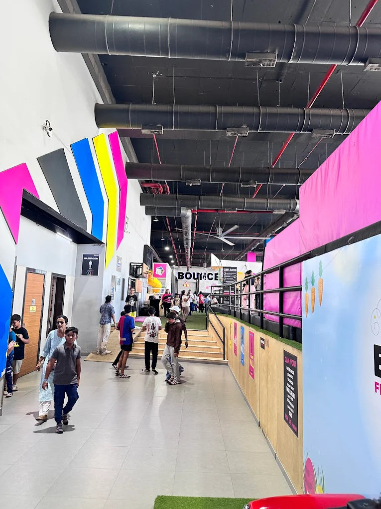 People walk through a brightly colored indoor trampoline park with large pipes overhead.