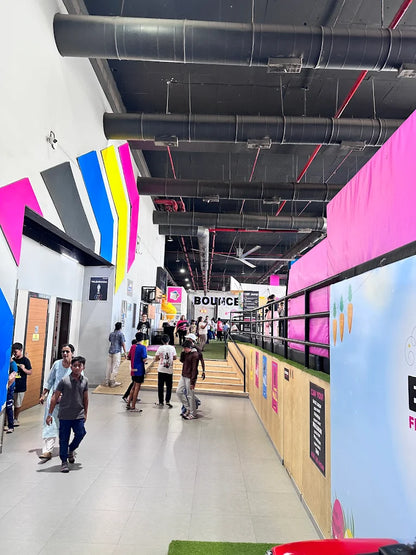People walk through a brightly colored indoor trampoline park with large pipes overhead.