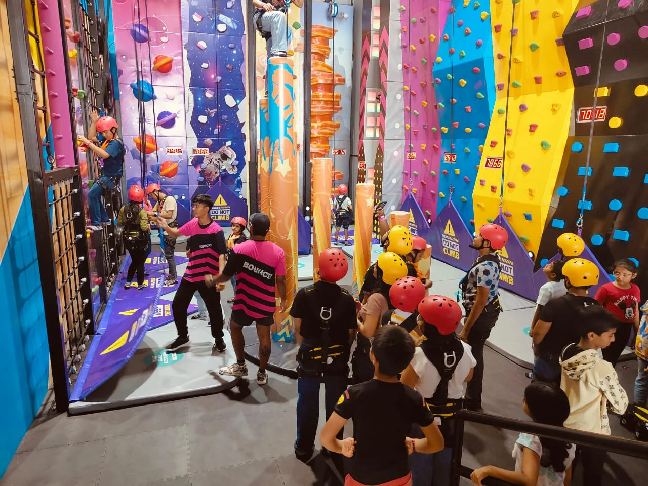 Children and adults in helmets climb colorful rock walls at an indoor adventure park.