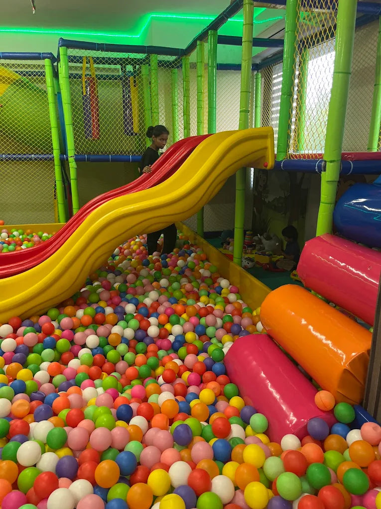 A young girl stands on a colorful ball pit slide in a children's indoor playground.