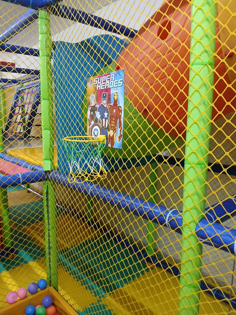 A colorful indoor playground with a superhero theme, featuring a basketball hoop and a ball pit for young children.