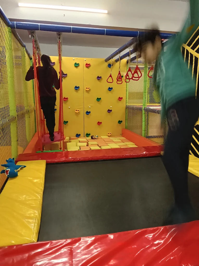 Children play in an indoor adventure park with a climbing wall and trampoline.