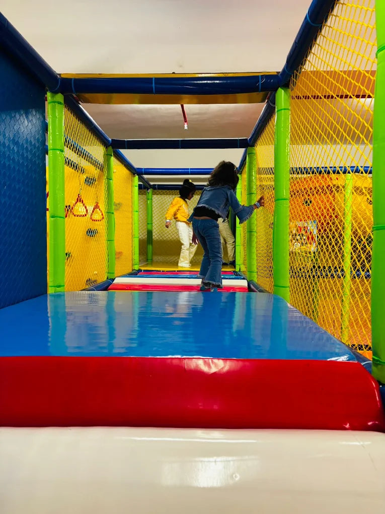 Children play in a colorful indoor playground with padded surfaces and netting.