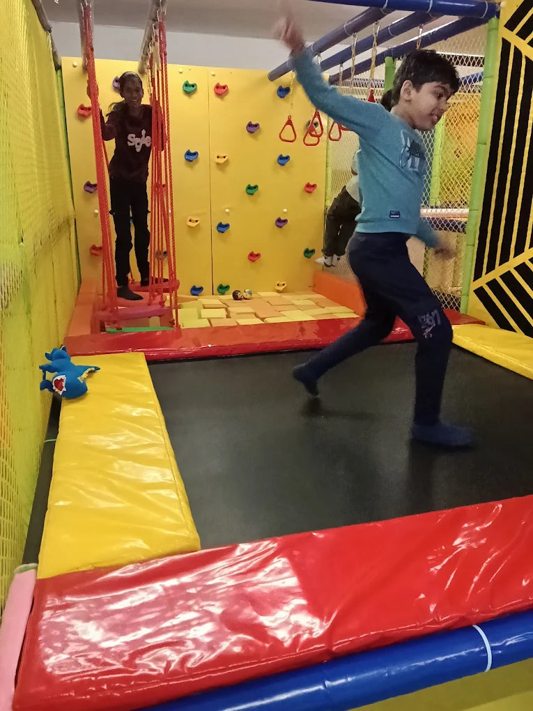 Two children play in an indoor adventure park with a climbing wall and trampoline.