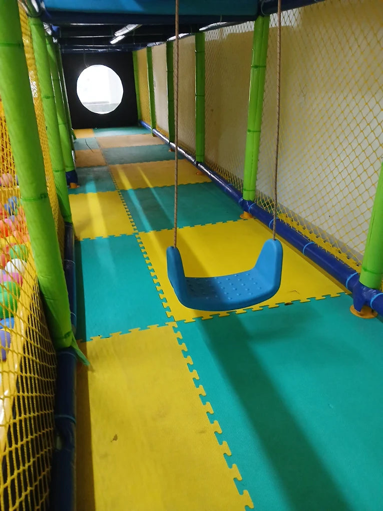 A blue swing hangs in a colorful indoor playground with a ball pit on the left. The floor is made of interlocking foam mats in yellow and green.