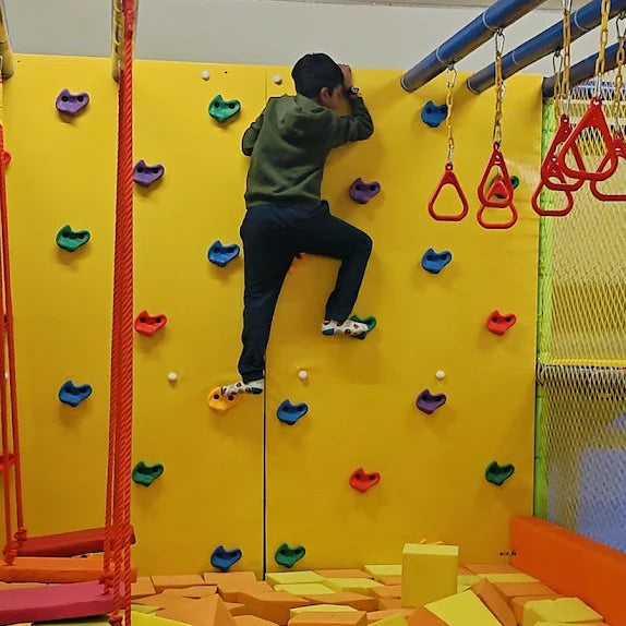 A young boy climbs a yellow indoor rock-climbing wall with colorful handholds.