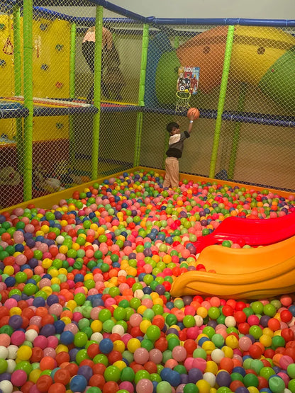 A young child plays basketball in a colorful ball pit at an indoor playground.