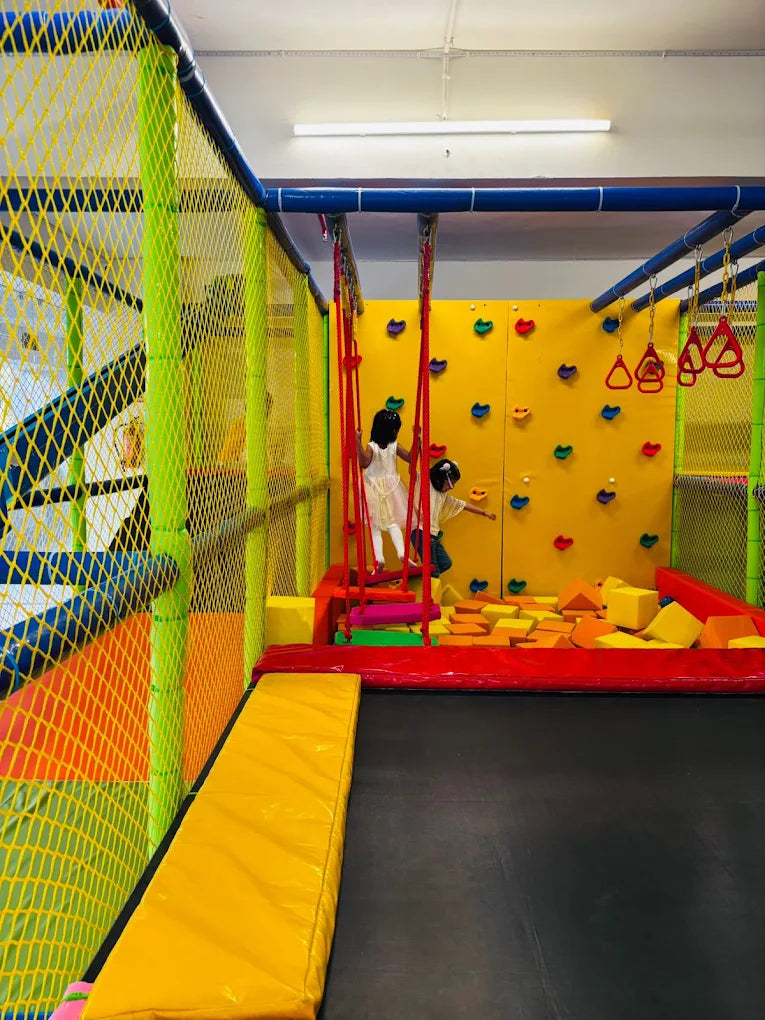 Children play in a colorful indoor playground with a trampoline, foam pit, and climbing wall.