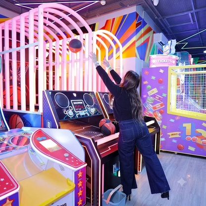 A young woman plays a basketball arcade game in a brightly lit entertainment center.