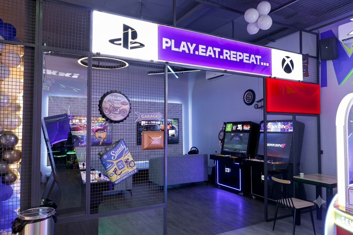A brightly lit gaming lounge with arcade machines, PlayStation and Xbox logos, and a "Play. Eat. Repeat." sign.