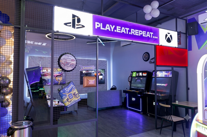 A brightly lit gaming lounge with arcade machines, PlayStation and Xbox logos, and a "Play. Eat. Repeat." sign.