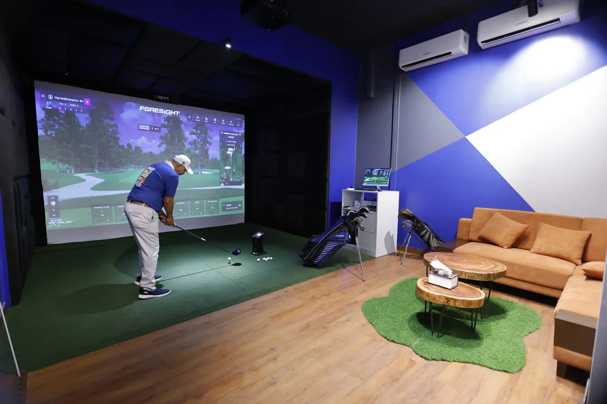 A man practices golf on a simulator in a room with a couch and golf bags.