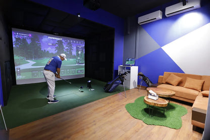 A man practices golf on a simulator in a room with a couch and golf bags.