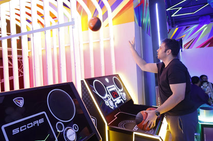 A man plays a basketball arcade game with neon lights and a digital scoreboard.