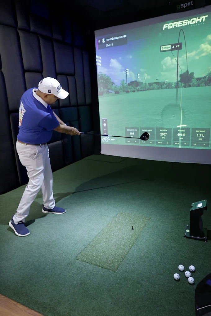 A man in a blue shirt and white hat swings a golf club in a simulator room. The screen shows a golf course with shot data.