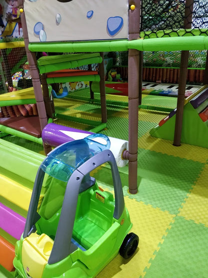 A green toy car sits on a colorful play mat in a children's indoor playground. The playground features slides and climbing structures.
