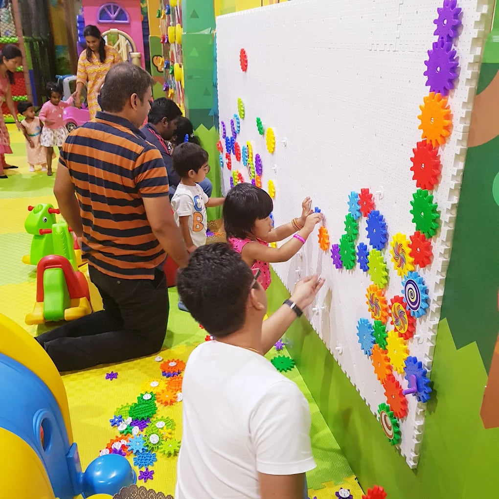 Children and adults play with colorful gears on a large white wall in an indoor play area.