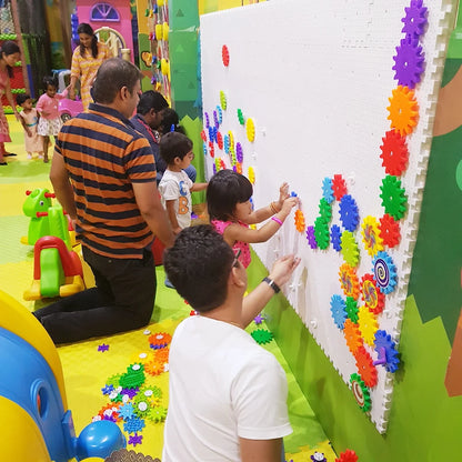 Children and adults play with colorful gears on a large white wall in an indoor play area.