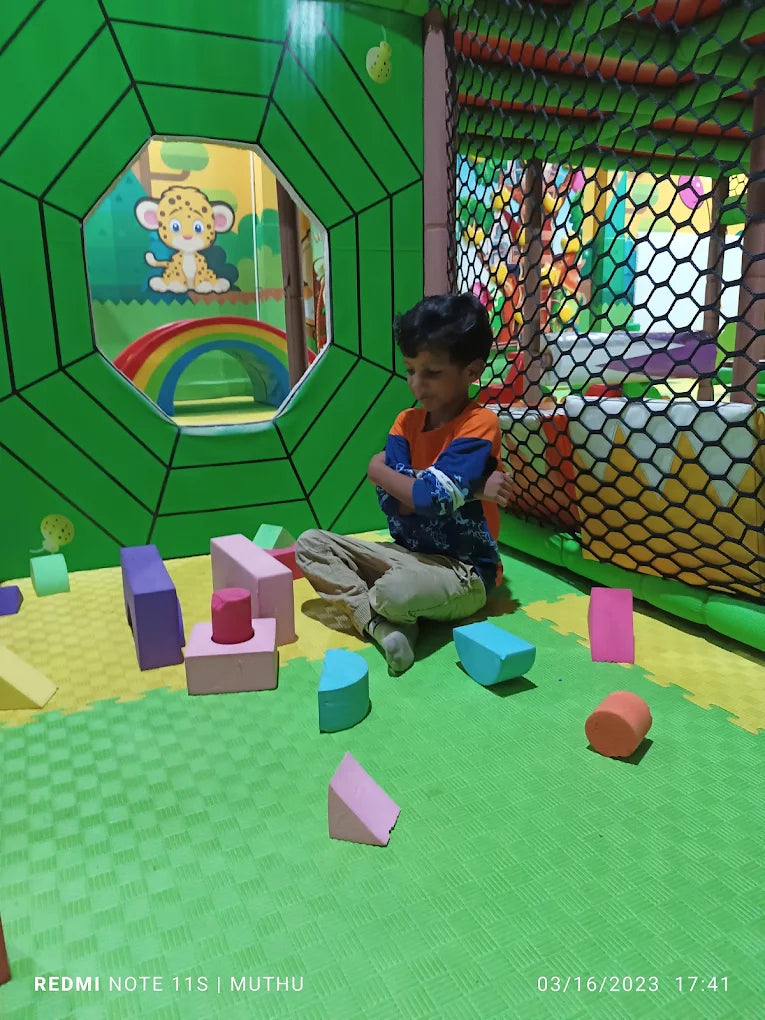 A young boy sits with his arms crossed in a colorful indoor playground, surrounded by soft building blocks.