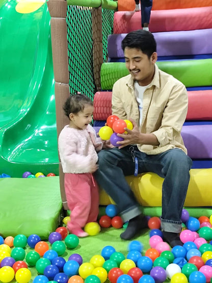 A father and toddler play with colorful balls in a ball pit at an indoor playground.