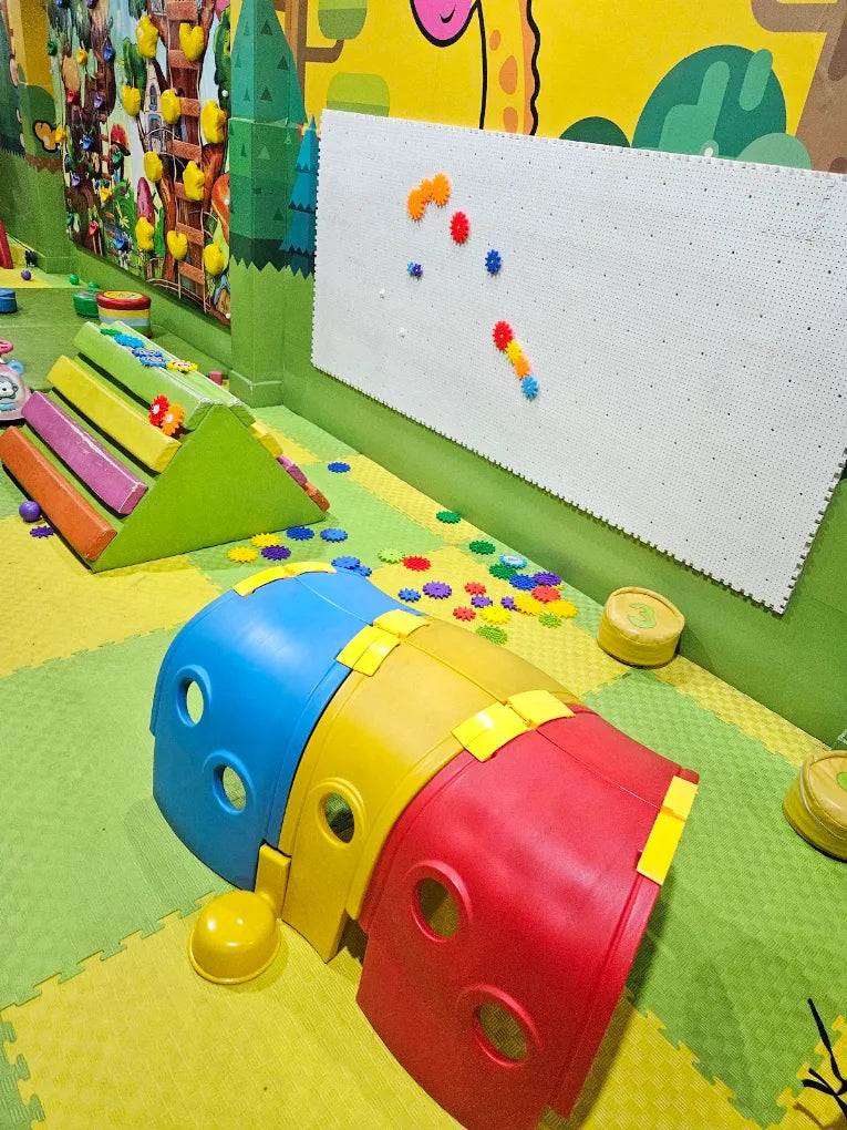 A colorful indoor play area for young children with a climbing structure, tunnel, and magnetic board.