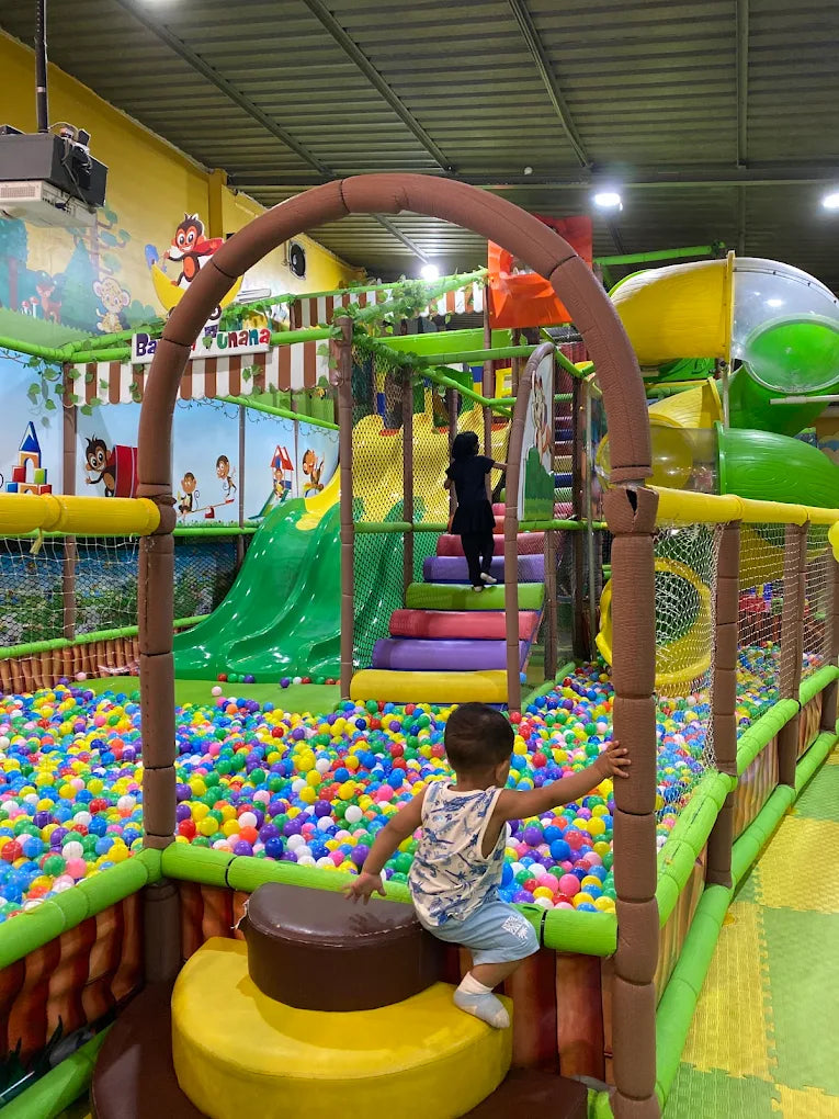 A young child sits on a play structure overlooking a ball pit in an indoor playground.