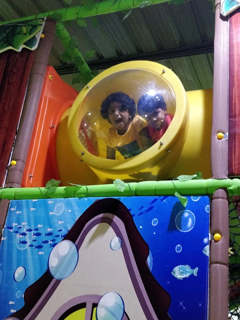 Two young children peek out of a yellow tube in a colorful indoor playground.