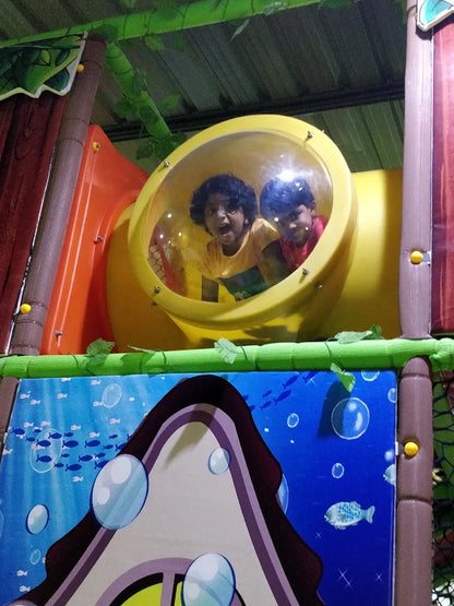 Two young children peek out of a yellow tube in a colorful indoor playground.