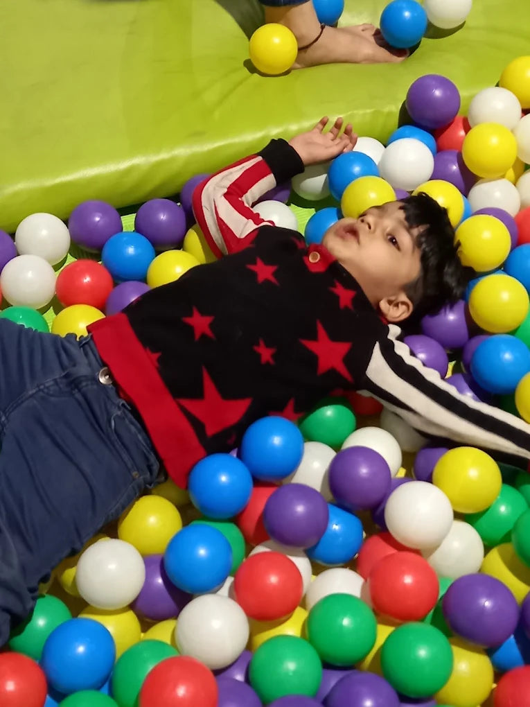 A young child lies in a ball pit, surrounded by colorful balls.