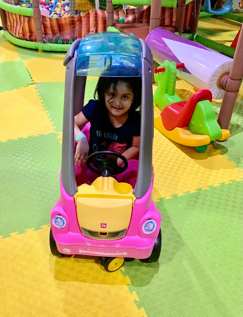 A young girl sits in a pink toy car at an indoor playground.