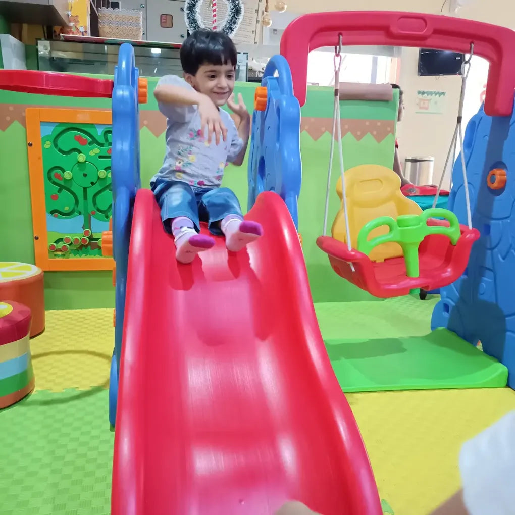A young child slides down a red slide in a colorful indoor playground.