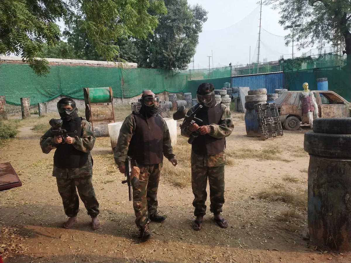 Three young adults in camouflage gear and masks stand ready for a paintball game in an outdoor arena.
