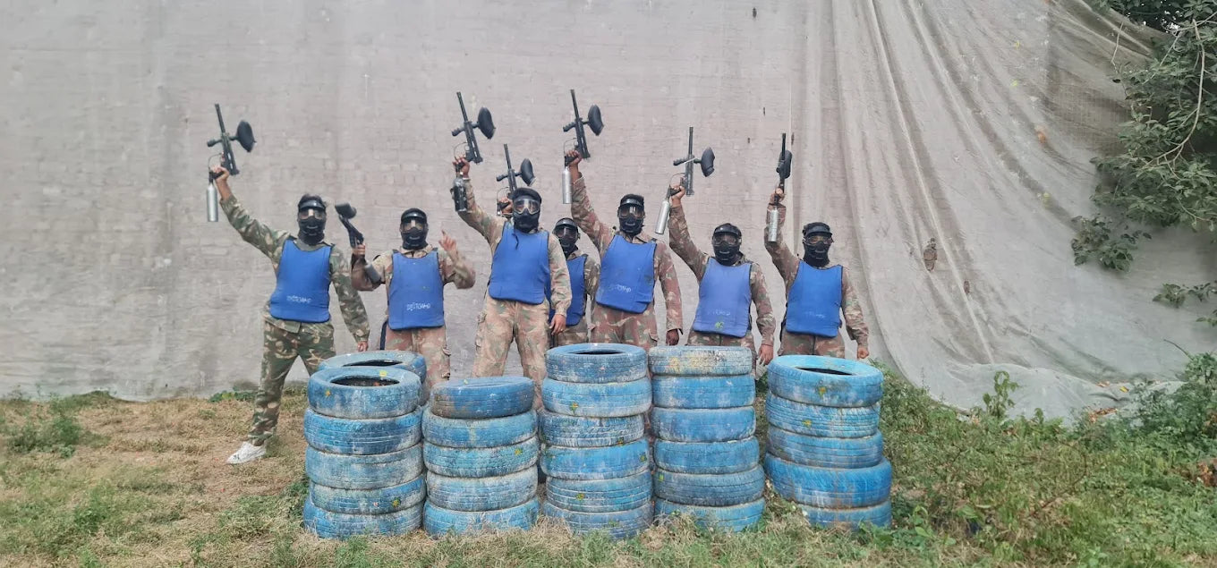 A group of young adults in paintball gear and masks pose with their paintball guns raised in front of a concrete wall and tarp.