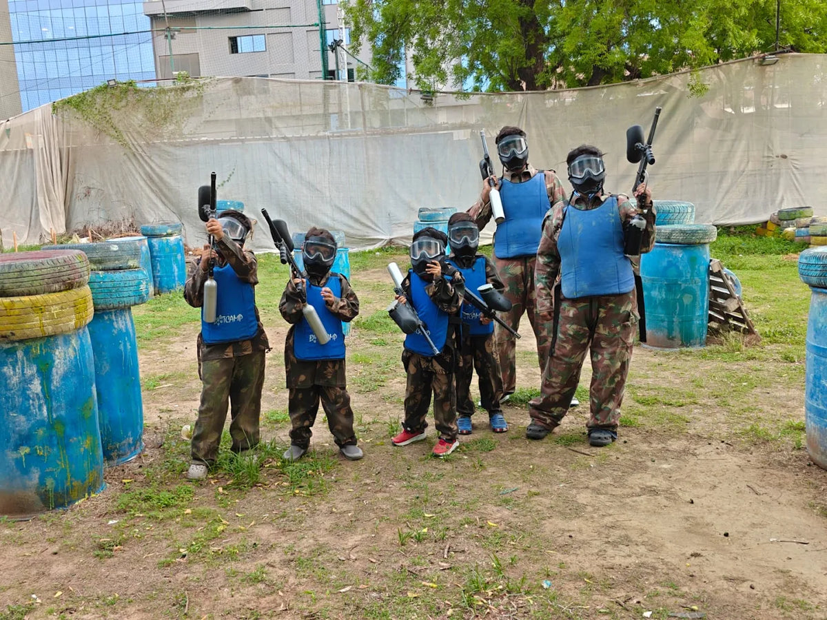 Children in paintball gear stand ready with their markers at an outdoor paintball field.