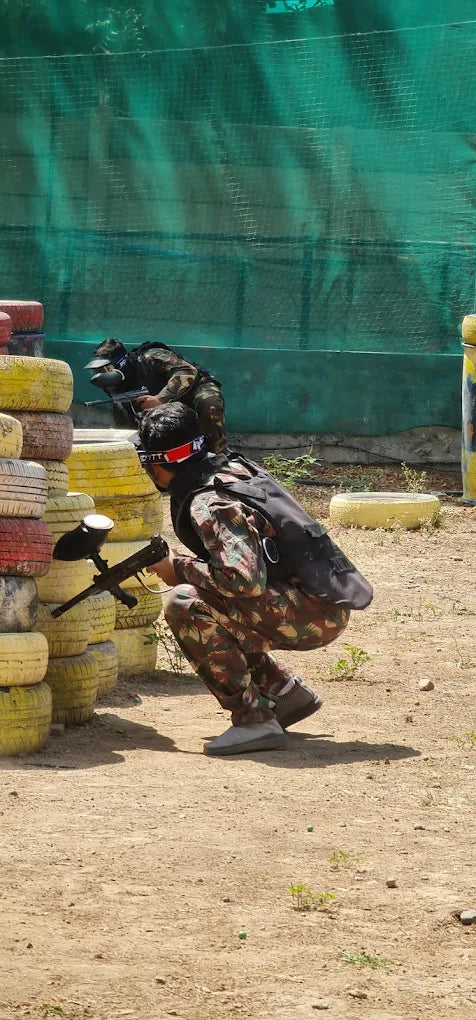 Two young people in camouflage gear play paintball, crouching behind tire barriers.