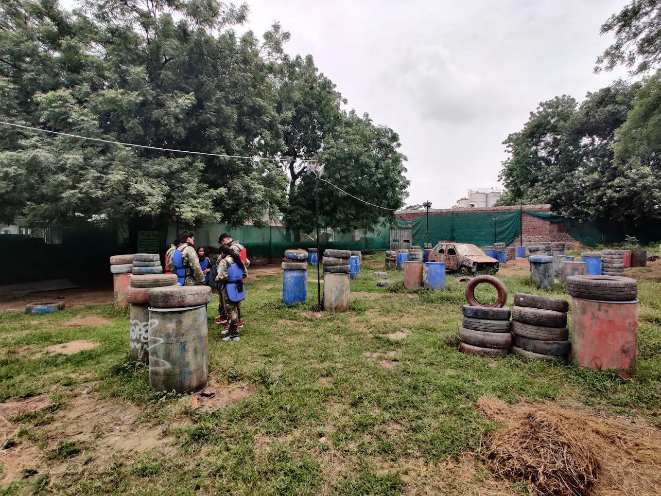 Young adults in protective gear gather at a paintball field with barrels and a derelict car as obstacles.