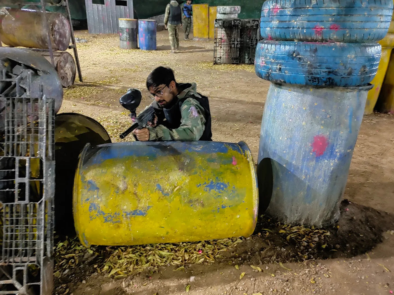 A young man plays paintball, taking cover behind a yellow barrel. The paintball field is littered with debris and obstacles.