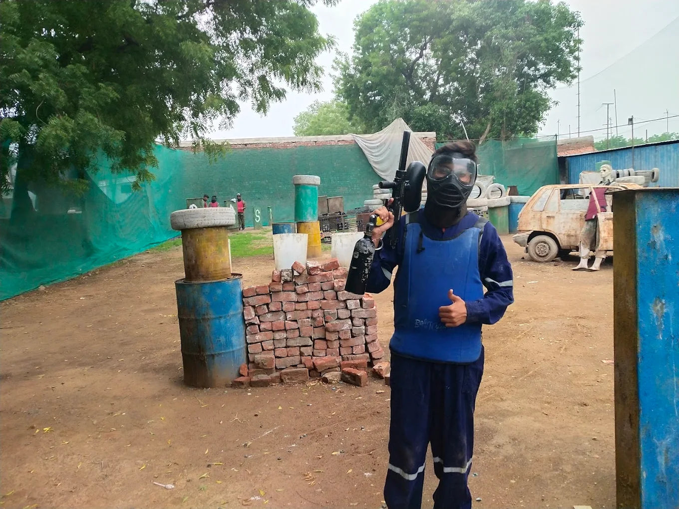 A young person in paintball gear poses with a marker, ready for a game on an outdoor course.