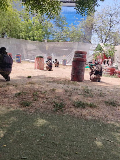 Adults playing paintball in an outdoor arena with barrels and netting.
