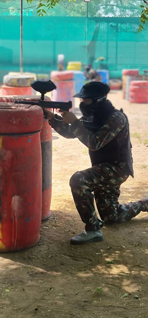 A person in camouflage kneels behind a red barrel, aiming a paintball gun. They are wearing a mask and helmet in an outdoor paintball arena.